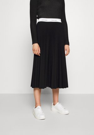 Kobieta ubrana w czarny prążkowany top, czarną plisowaną spódnicę midi z białym paskiem Calvin Klein oraz białe sznurowane trampki, stojąca na szarej podłodze.