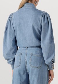 Lichtblauwe denim top met opgeblazen mouwen, knoopmanchetten en een hoge kraag. Heeft een soepele textuur en subtiele stikseldetails.