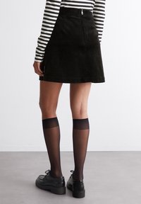 Gonna a trapezio nera in corduroy con zip sul retro, abbinata a calze nere fino al ginocchio e scarpe nere con lacci. Camicia a maniche lunghe a righe visibile.