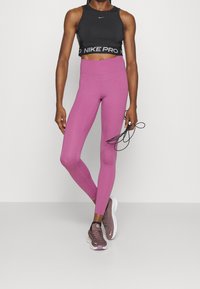 Femme en haut court noir et leggings roses tenant une corde à sauter noire, portant des baskets violettes, debout devant un fond blanc uni.