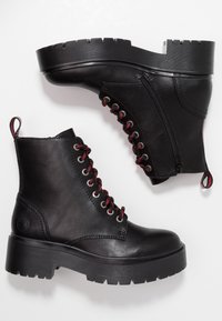 Botas de cuero negro hasta el tobillo con una suela gruesa, featuring cordones rojos, cremallera lateral y tira para calzar. Parte superior texturizada y suela con agarre.