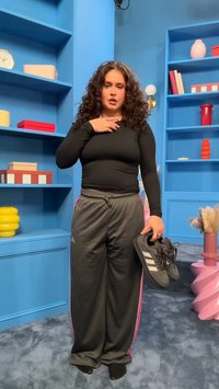 Femme aux cheveux bouclés portant un haut noir et un pantalon gris tient des baskets noires dans une pièce bleue avec des étagères et des objets décoratifs.