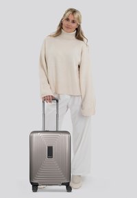 Valigia da cabina in grigio metallico con superficie rigata, manico retrattile e quattro ruote, abbinata a un maglione crema e pantaloni bianchi larghi.