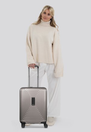 Valigia da cabina in grigio metallico con superficie rigata, manico retrattile e quattro ruote, abbinata a un maglione crema e pantaloni bianchi larghi.