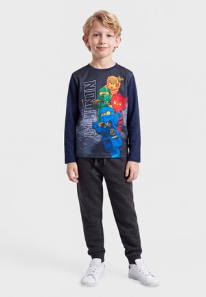 Jeune garçon debout portant un t-shirt à manches longues graphique Ninjago bleu marine, un pantalon de jogging noir et des baskets blanches sur fond blanc.
