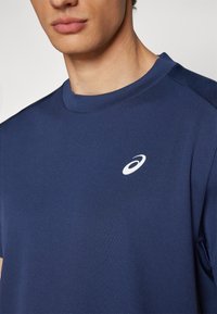 Marineblaues Athletic-T-Shirt mit Rundhalsausschnitt, kurzen Ärmeln und einem weißen Logo auf der Brust. Glatte Textur mit einem dezenten Stoffmuster.