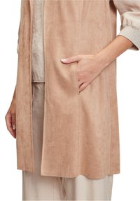 Jasnoróżowa kamizelka z zamszu z otwartym przodem, boczną kieszenią i gładką fakturą, noszona na jasnobeżowej bluzce.