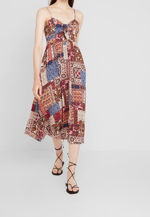 Frau trägt ein ärmelloses Midikleid mit Patchwork-Muster, dünnen Trägern und einer vorderen Schleife, kombiniert mit schwarzen, ristrigen Sandalen.