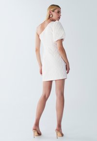 Bijela, kviltana mini haljina s jednim off-shoulder rukavom i napuhanim detaljima; uparena s bež štiklama.