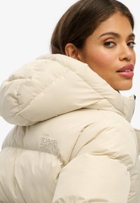 Veste beige rembourrée avec une capuche, présentant un design matelassé et un logo "Superdry" en relief sur l'épaule gauche. La texture semble lisse.