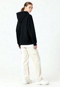 Felpa nera oversize con cappuccio, abbinata a pantaloni cargo color crema con due tasche laterali e scarpe da ginnastica bianche chunky.