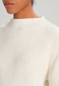 Cremfarbener Strickpullover mit hohem Kragen, geripptem Muster und lässiger Passform. Das Material wirkt weich und leicht.
