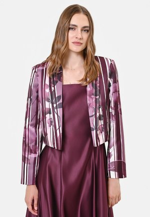 Jeune femme aux longs cheveux châtain clair portant une robe en satin violette sans manches et une veste courte rayée florale aux tons violets.