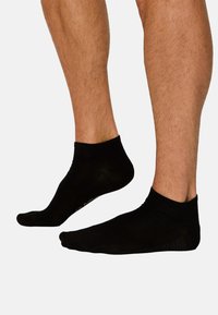 Zwei behaarte Unterschenkel mit schwarzen Knöchelsocken, ein Fuß leicht vom Boden abgehoben vor einem schlichten hellen Hintergrund.