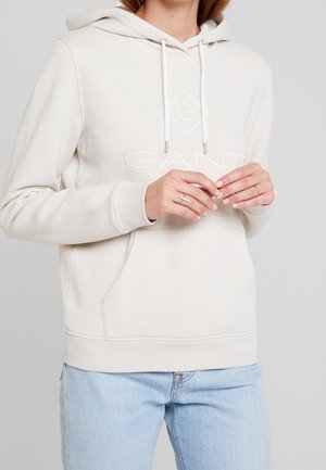 Personne portant un sweat à capuche GANT beige clair avec cordons et poche avant, associé à un jean bleu clair, mains légèrement jointes.
