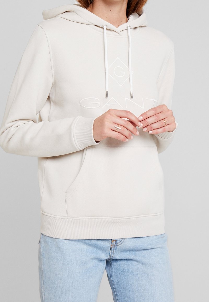 Personne portant un sweat à capuche GANT beige clair avec cordons et poche avant, associé à un jean bleu clair, mains légèrement jointes.