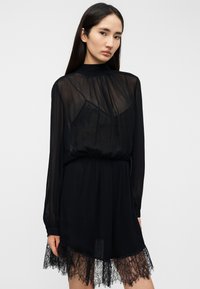 Μαύρο φόρεμα με διαφανή μακριά μανίκια και ψηλό λαιμό. Διαθέτει εφαρμοστή μέση και δαντέλα στο τελείωμα, που αναδεικνύει ένα λεπτό σχέδιο.