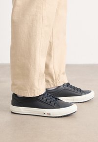 Schwarze Leder-Sneaker mit weißen Gummisohlen, versehen mit Schnürsenkeln und einer strukturierten Oberfläche. Akzentuiert mit einem kleinen detail in Rot, Weiß und Blau.