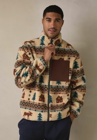 Kurtka polarowa zapinana na zamek z kremowym tłem, ozdobiona wzorem z łosi, sosnami i chatkami w kolorach brązowym i turkusowym. Posiada brązową kieszeń na piersi.