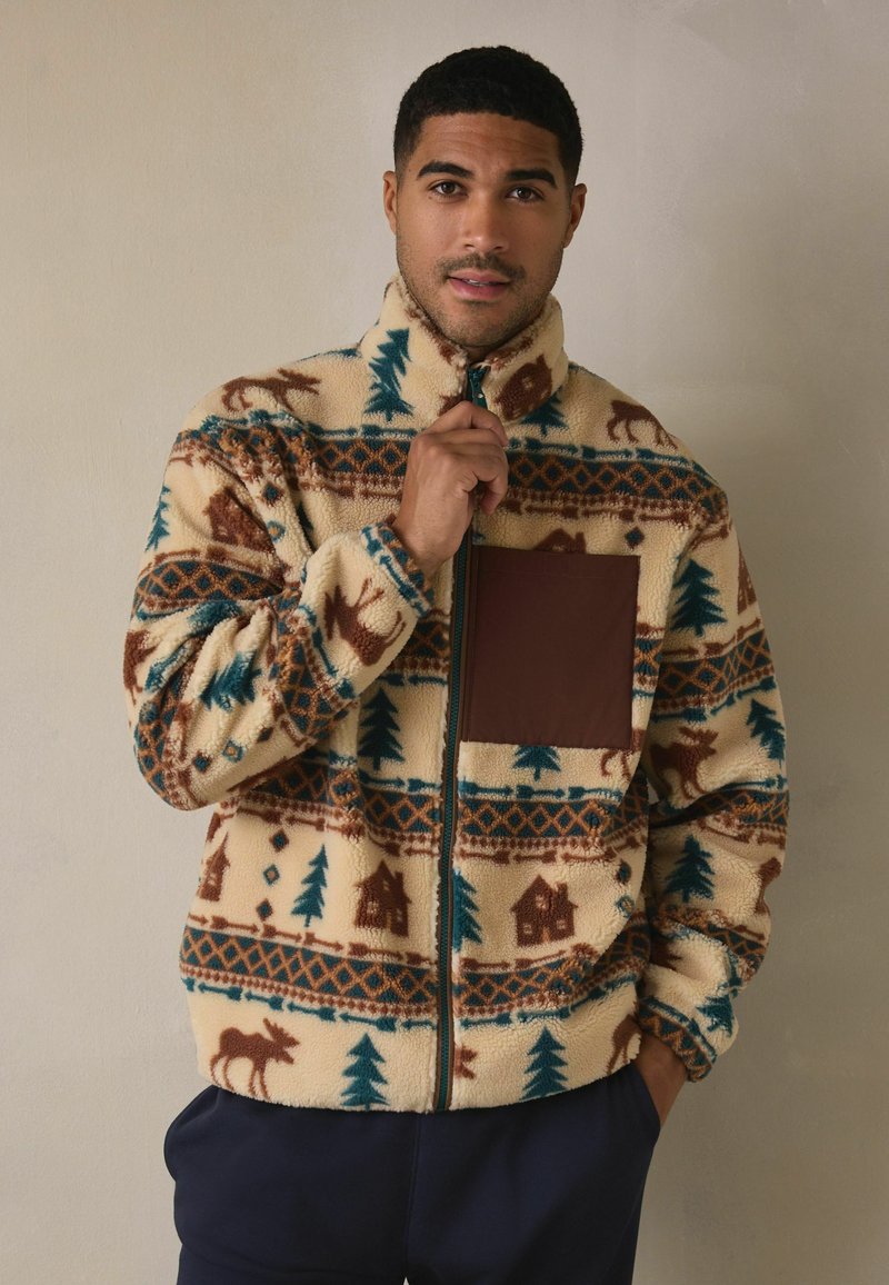 Kurtka polarowa zapinana na zamek z kremowym tłem, ozdobiona wzorem z łosi, sosnami i chatkami w kolorach brązowym i turkusowym. Posiada brązową kieszeń na piersi.