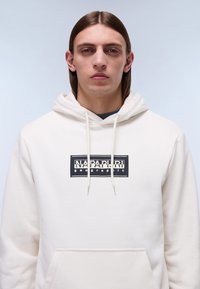 Sudadera con capucha color crema, con un bolsillo frontal y un logotipo rectangular negro. Textura suave con cordones en la capucha.