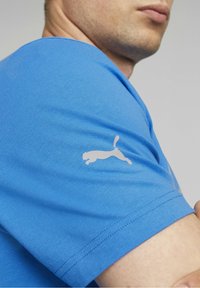 Blå bomullströja med korta ärmar, med en reflekterande vit Puma-logga på vänster ärm och rund halsringning. Mjuk textur.