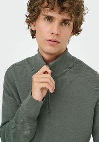 Sudadera de media cremallera de color verde con canalé, fabricada en un tejido texturizado, que cuenta con un cuello alto y detalle de cremallera. Mangas y escote ajustados.