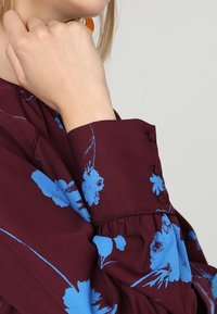 Blouse bordeaux avec motif floral bleu, détail de manche froncée et poignets boutonnés. Tissu doux avec un drapé et une texture visibles.