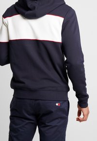 Felpa blu navy con una striscia orizzontale bianca e dettagli rossi, realizzata in un misto cotone. Presenta una tasca a marsupio e polsini a coste.
