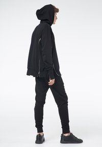 Czarna bluza z kapturem z zamkiem, połączona z czarnymi joggerami. Materiał wydaje się być miękki, charakteryzuje się luźnym krojem i ściągaczami przy rękawach. Czarny obuwie sportowe dopełnia stylizację.