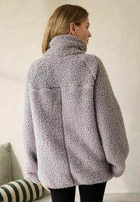 Grå, fuzzy oversized fleecejakke med høj krave og sænkede skuldre. Har et enkelt design og tekstureret stof for at give varme.