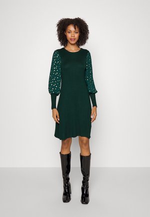 Robe en tricot verte avec de longues manches bouffantes ornées de pois dorés. Texture lisse, coupe à hauteur du genou, assortie à des bottes noires brillantes montantes jusqu'au genou.