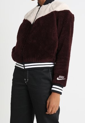 Osoba nosząca bordowo-białą puszystą kurtkę Nike z paskowanymi mankietami i dołem, w połączeniu z czarnymi spodniami.