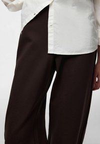 Bijela košulja s dugim rukavima, djelomično uvučena u široke tamnobraon hlače, s glatkom teksturom i minimalističkim dizajnom s čistim linijama.