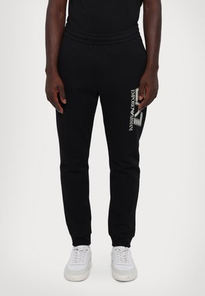 Homme portant un pantalon de survêtement noir avec le logo blanc "Emporio Armani" sur la cuisse droite et des baskets blanches, debout devant un fond uni.