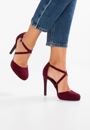 Chaussures à talons hauts bordeaux en daim, avec un bout ouvert et des lanières croisées à la cheville. Le talon est fin et élégant.