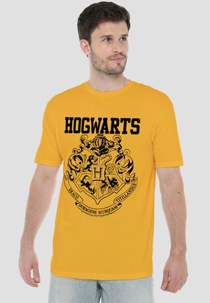 Mann trägt gelbes T-Shirt mit Hogwarts-Wappen und Motto, steht vor einem einfarbigen hellen Hintergrund und blickt seitlich.