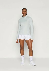 Helles graues Langarmoberteil mit einem Viertel-Reißverschluss, kombiniert mit weißen Sportshorts. Glattes Material, minimalistisches Design und ein dezentes Logodetail.