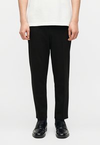 Πρόσθια όψη ατόμου που φοράει μαύρο παντελόνι ραμμένο κατά παραγγελία, μαύρα μοκασίνια και λευκό πουκάμισο, στέκεται μπροστά σε απλό φωτεινό φόντο.