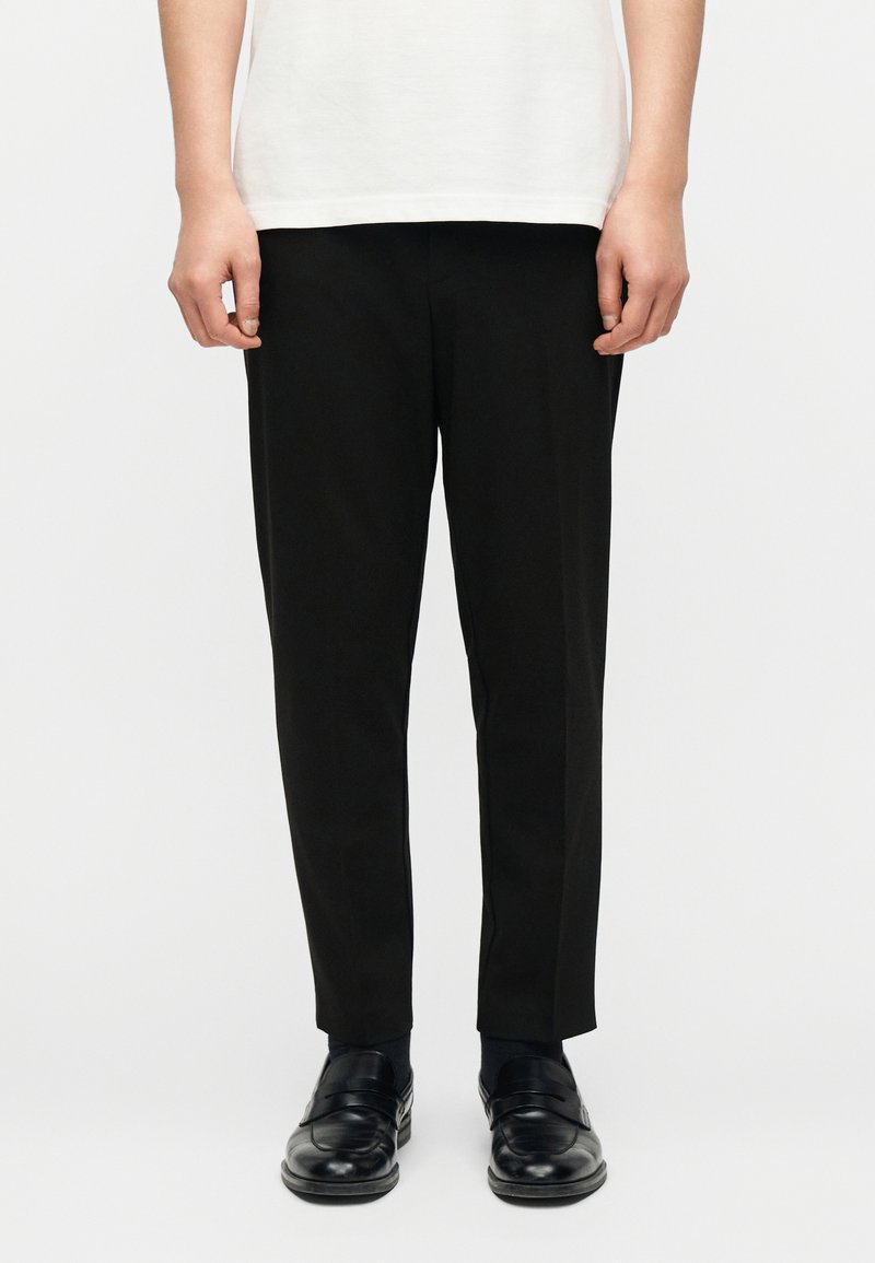 Πρόσθια όψη ατόμου που φοράει μαύρο παντελόνι ραμμένο κατά παραγγελία, μαύρα μοκασίνια και λευκό πουκάμισο, στέκεται μπροστά σε απλό φωτεινό φόντο.