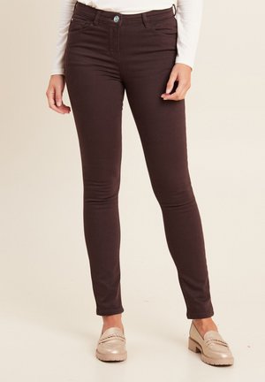 Bruine skinny jeans met een gladde textuur, hoge tailleband en voorzakken. Het model draagt lichtgekleurde instappers voor contrast.