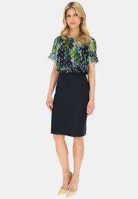 Kwiatowa przezroczysta bluzka w odcieniach zieleni i niebieskiego na ciemnym tle, zestawiona z dopasowaną granatową spódnicą ołówkową i beżowymi szpilkami z paskiem na kostce.