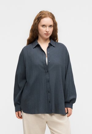 Frau mit langen roten Haaren, die ein locker sitzendes dunkelblaues Hemd mit Knöpfen und hellbeige Hose trägt, steht vor einem schlichten weißen Hintergrund.