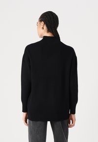 Fekete kötött pulóver magas nyakkal, laza fazonnal, hosszú ujjal, bordás mandzsettával és szegéllyel. Simaság jellemzi semleges háttérrel.