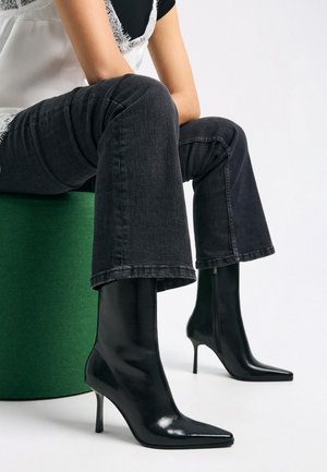 Fekete, fényes magas sarkú bokacsizma hegyes orrformával, sötét farmernadrággal párosítva, zöld hengeres tárgyon ülve.