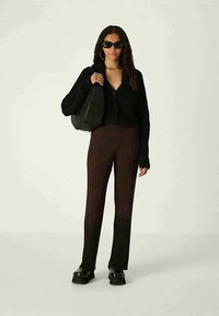 Cárdigan negro de botones, pantalones marrones de canalé, bolso negro grande y zapatos de plataforma negros. Los accesorios incluyen gafas de sol grandes y pendientes de aro.