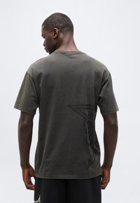 Άνδρας με κοντά μαλλιά που φοράει ένα σκούρο ξεθωριασμένο μπλουζάκι με μια μεγάλη μαύρη αστέρι στο κάτω δεξί μέρος της πλάτης, στέκεται μπροστά σε απλό φωτεινό φόντο.