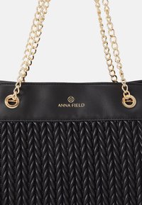 Bolso negro con un patrón tejido texturizado. Presenta una correa de cadena en tono dorado y acentos metálicos. Logo de la marca exhibido de forma prominente.