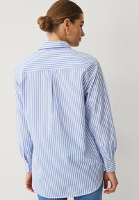 Camisa de rayas azul y blanca con mangas largas, puños abotonados, yugo trasero y detalle de pliegue. Fabricada con tejido ligero, corte relajado.