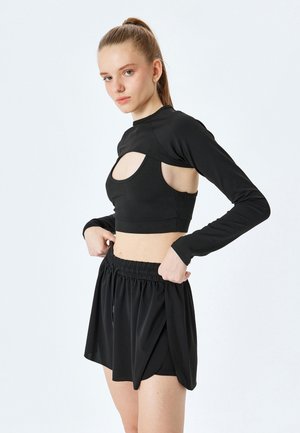 Musta urheilu crop-top, jossa on pitkät hihat ja leikkauksen yksityiskohta, yhdistettynä mustiin, pleatattuihin, elastisella vyötäröllä varustettuihin shortseihin. Sujuva pinta.